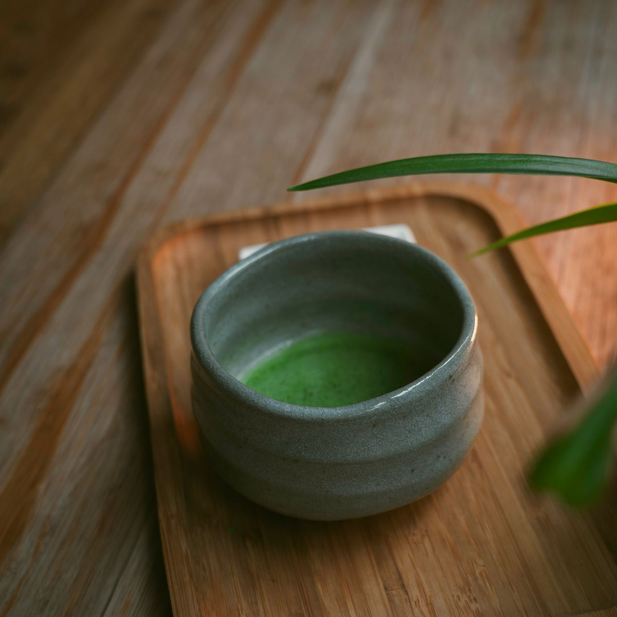 A ceramic bowl filled with matcha green tea, set on a wooden tray indoors.
