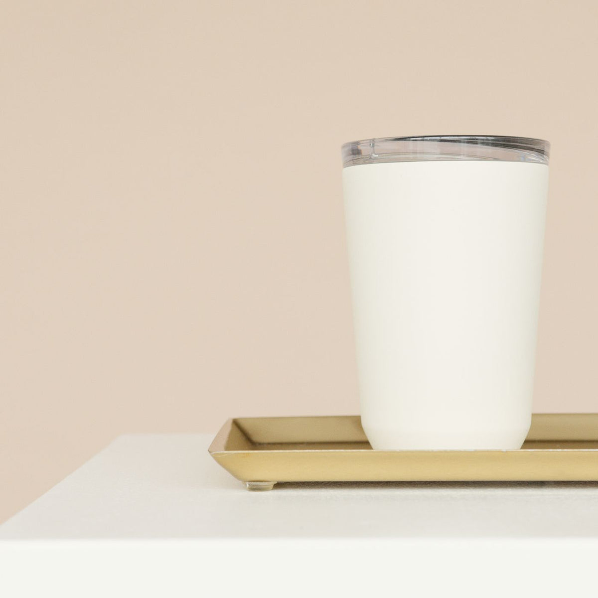 A white tumbler standing on a gold tray against a beige background.